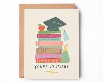 Puede incluir: Una tarjeta de felicitación con una pila de libros coloridos coronada por un birrete gris oscuro y una manzana roja. Un portalápices verde con dos lápices amarillos se sitúa al lado. El texto dice YOU'RE SO SMART con un lápiz amarillo debajo.