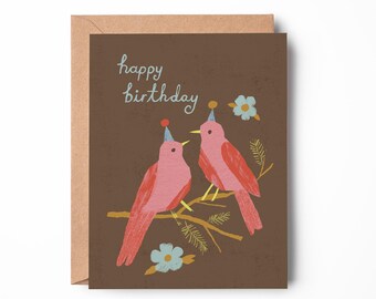 May include: A greeting card with a brown background and a brown envelope. The card features two pink birds wearing party hats perched on a branch. The words "happy birthday" are written in light blue.