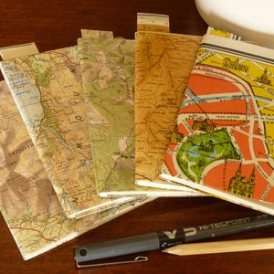 May include: A stack of six small, vintage, paperbound books with maps printed on the covers. The maps are in various colours and depict different locations. The books are arranged in a row on a wooden surface. A black pen and a wooden pencil are lying next to the books.