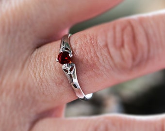 May include: A silver ring with a small, round, red gemstone set in the center. The ring has a delicate, openwork design on the band, with a floral or leaf-like pattern. The ring is on a finger with pink nail polish.