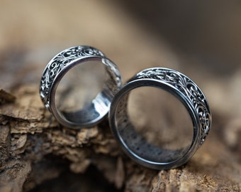 May include: Two silver rings with intricate floral designs. The rings are stacked on top of each other and are resting on a piece of weathered wood.