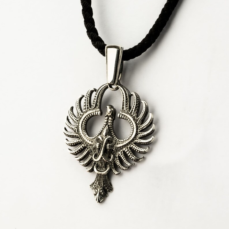 May include: A silver pendant necklace with a detailed design of a phoenix with its wings spread. The pendant is attached to a black cord.