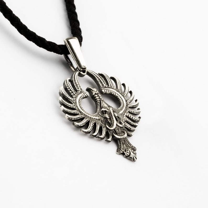 May include: A silver pendant necklace featuring a detailed, intricate design of a stylized phoenix with outstretched wings. The pendant is attached to a black cord.