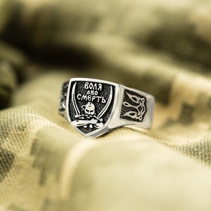 May include: A silver ring with a black shield-shaped design featuring a skull and crossbones and the text "ВОЛЯ АБО СМЕРТЬ" in Cyrillic script. The ring has a textured band with a stylized design.