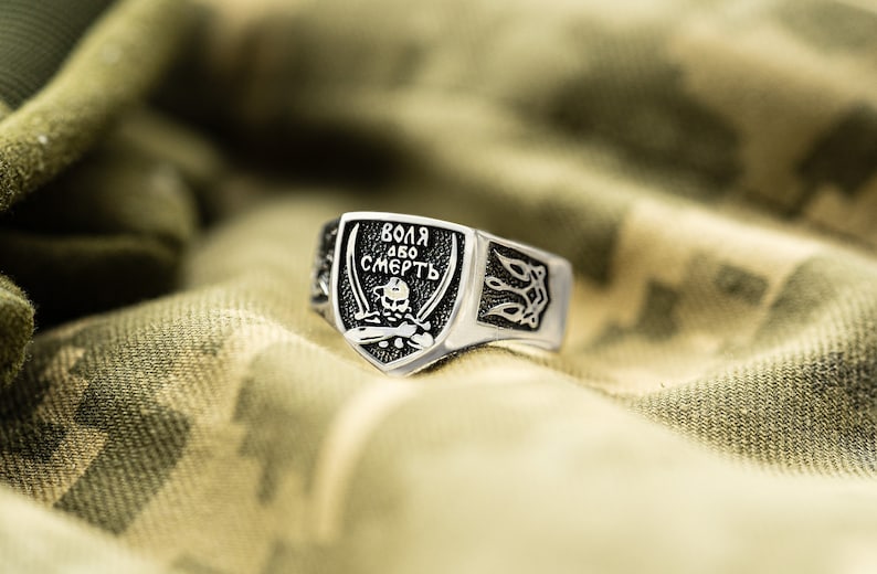May include: A silver ring with a black shield-shaped design featuring a skull and crossbones and the text "ВОЛЯ АБО СМЕРТЬ" in Cyrillic script. The ring has a textured band with a stylized design.
