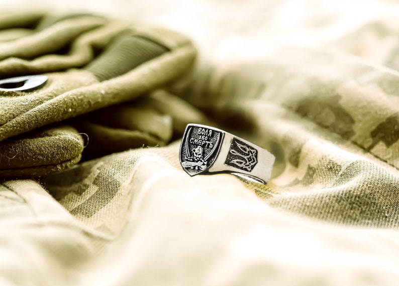 May include: A silver ring with a shield-shaped design featuring a skull and crossbones, the inscription "ВОЛЯ  ЛЕБО  СМЕРТЬ" and a trident. The ring is resting on a camouflage fabric.