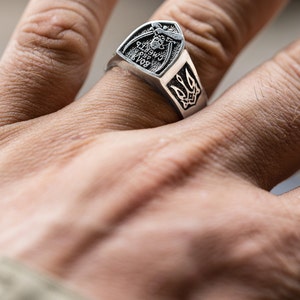 May include: A silver ring with a shield-shaped design featuring a trident and the text "Glory to Ukraine".