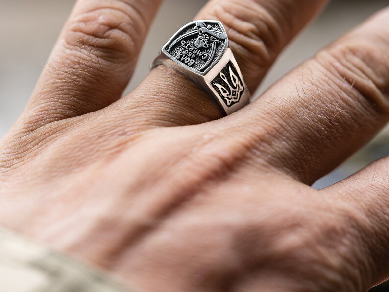 May include: A silver ring with a shield-shaped design featuring a trident and the text "Glory to Ukraine".