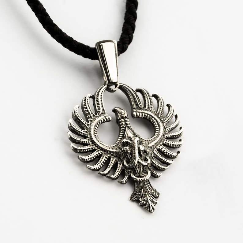 May include: A silver pendant necklace featuring a detailed design of a phoenix with outstretched wings. The pendant is attached to a black cord.