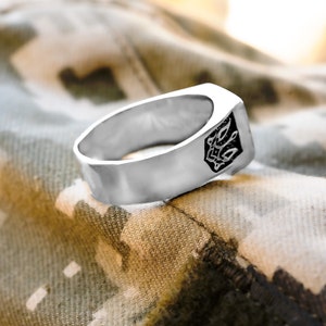 May include: A silver ring with a black design on the top. The ring is sitting on a camouflage fabric.