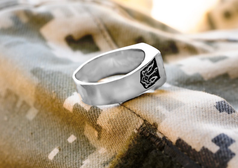 May include: A silver ring with a black design on the top. The ring is sitting on a camouflage fabric.