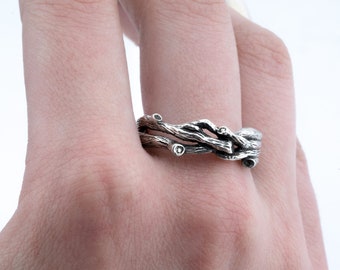 Può includere: Un anello d'argento con un designo di rami intrecciati e piccole pietre chiare.