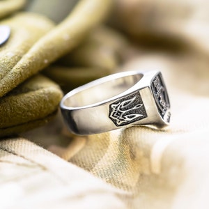 May include: A silver ring with a detailed design on the top. The ring is sitting on a beige fabric surface.
