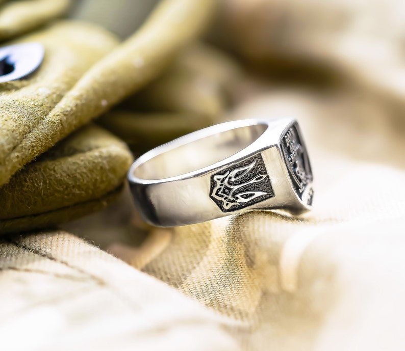May include: A silver ring with a detailed design on the top. The ring is sitting on a beige fabric surface.