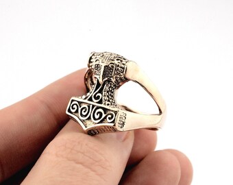 Puede incluir: Un anillo de color bronce con un diseño detallado de martillo, con intrincados patrones y una banda curva. La cabeza del martillo tiene forma cuadrada con detalles en espiral. El anillo está sostenido por un dedo.