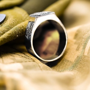 May include: A silver ring with an intricate design on the band. The ring is lying on a camouflage fabric.