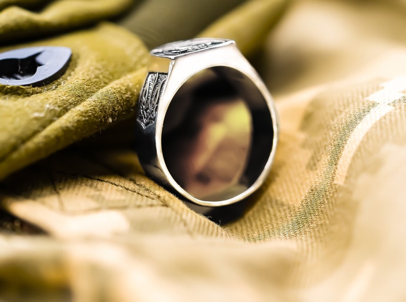 May include: A silver ring with an intricate design on the band. The ring is lying on a camouflage fabric.