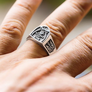 May include: A silver ring with a shield-shaped design featuring a trident and the inscription "Слава Україні, Героям Слава".