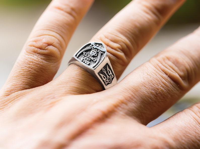 May include: A silver ring with a shield-shaped design featuring a trident and the inscription "Слава Україні, Героям Слава".