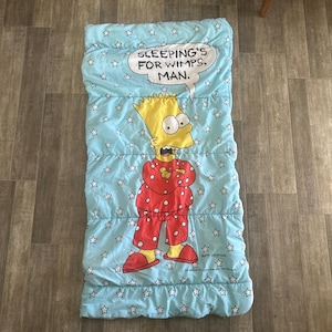 May include: A light blue sleeping bag featuring a cartoon image of Bart Simpson in red pajamas. The sleeping bag has a star pattern and a speech bubble that reads "SLEEPING'S FOR WIMPS, MAN."