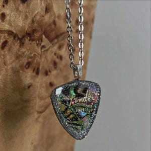 May include: Silver chain necklace with a guitar pick pendant. The pendant is a shimmering mix of colors with the word "Fender" and the word "THIN" visible. The background is a blurred wooden surface.