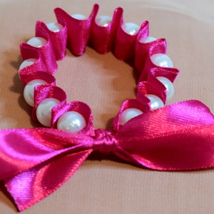 May include: A pink satin ribbon tied into a bow with a circle of white pearls and pink satin.