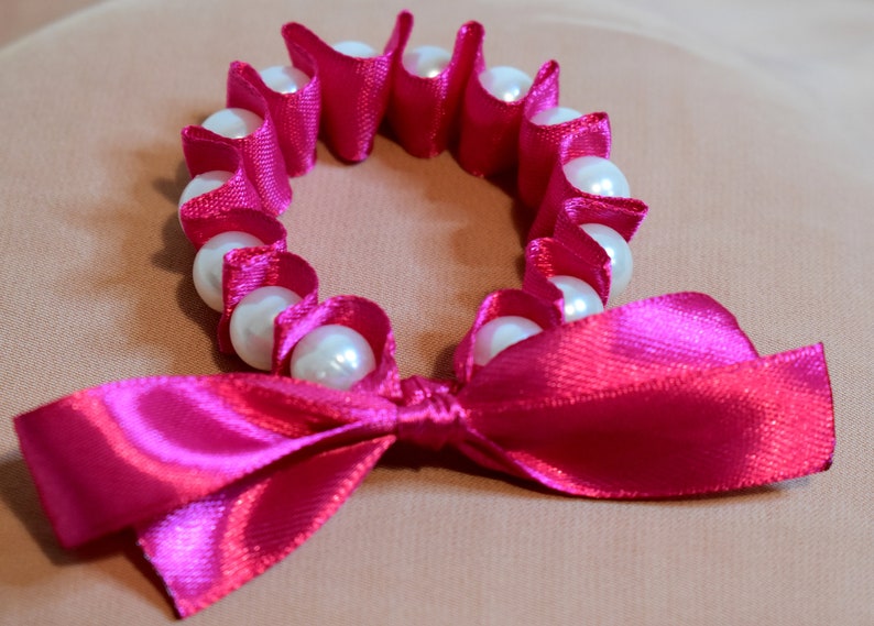 May include: A pink satin ribbon tied into a bow with a circle of white pearls and pink satin.