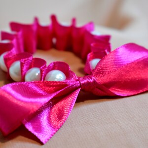 May include: A pink satin ribbon tied into a bow with a circle of white pearls.