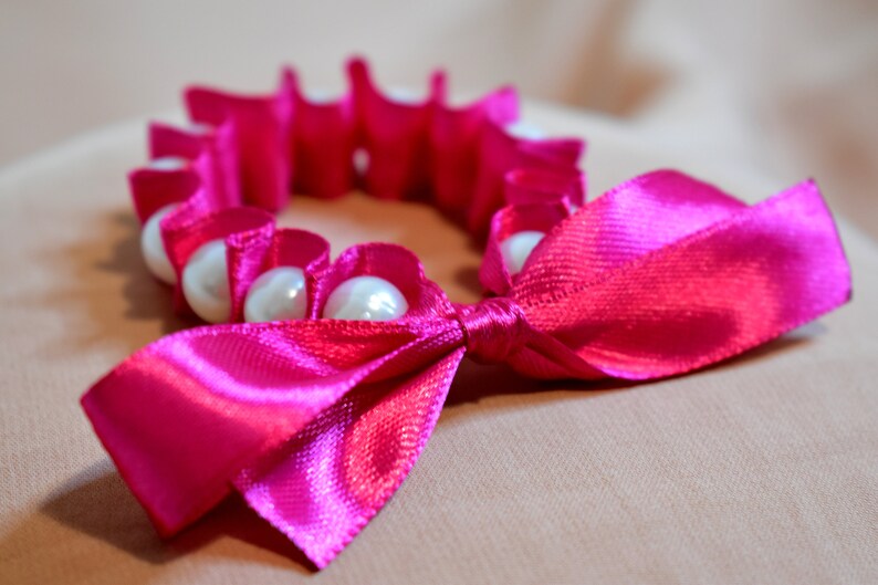 May include: A pink satin ribbon tied into a bow with a circle of white pearls.