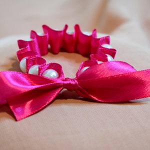May include: A pink satin ribbon tied into a bow with a circle of white pearl beads.