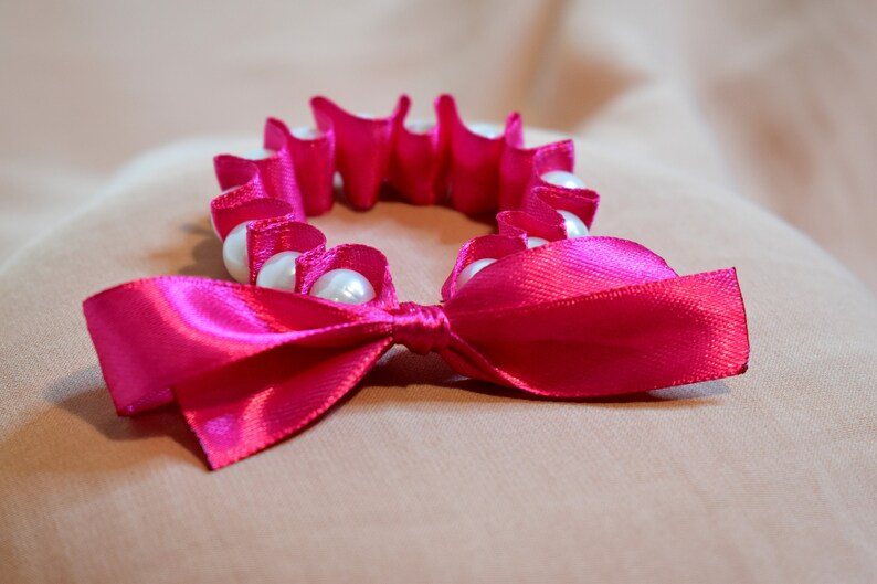 May include: A pink satin ribbon tied into a bow with a circle of white pearl beads.