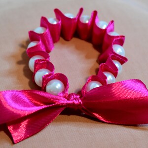 May include: A pink satin ribbon tied into a bow with a circle of white faux pearls strung on pink satin ribbon.