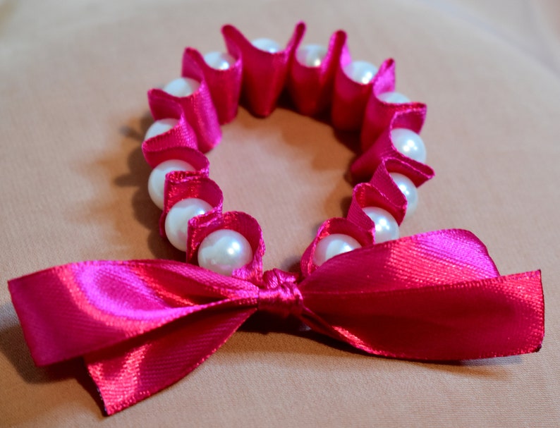 May include: A pink satin ribbon tied into a bow with a circle of white faux pearls strung on pink satin ribbon.