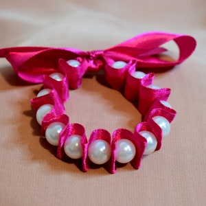 May include: A pink satin ribbon tied into a bow with a circle of white pearls strung through the ribbon.