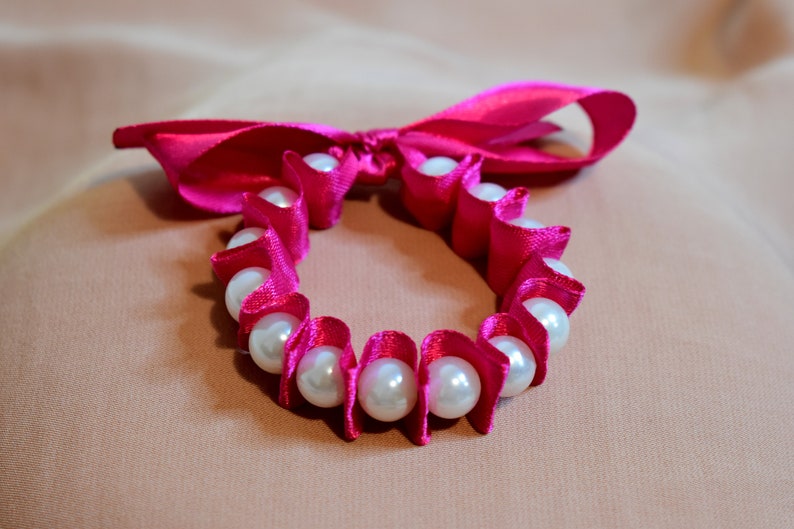 May include: A pink satin ribbon tied into a bow with a circle of white pearls strung through the ribbon.