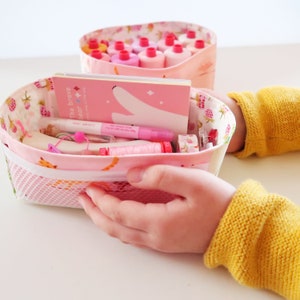 May include: Two pink fabric storage baskets with a white mesh bottom. The top basket is filled with pink spools of thread. The bottom basket is filled with craft supplies, including a pink pen, a white notepad with the text "The brave bear", and a pink and white sticker.