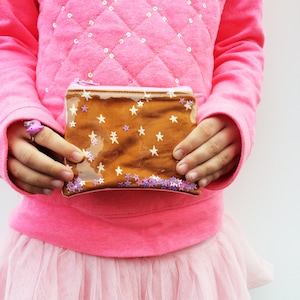 Peut inclure: Une petite pochette transparente de couleur marron avec une fermeture éclair. La pochette est décorée d'étoiles blanches et de paillettes violettes.