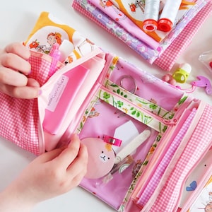 Può includere: Una pochette con cerniera in tessuto gingham rosa e bianco con una finestra in plastica trasparente. La pochette è aperta e piena di vari accessori per cucire, tra cui filo, un ago, un piccolo cerchio di stoffa rosa con un viso e un piccolo rettangolo di stoffa rosa con il testo "noma cocoon" stampato su di esso.