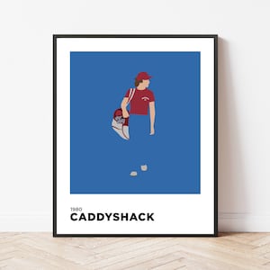 May include: A black framed poster with a blue background and a cartoon illustration of a person wearing a red hat and red shirt holding a golf bag. The text "1980 CADDYSHACK" is at the bottom of the poster.