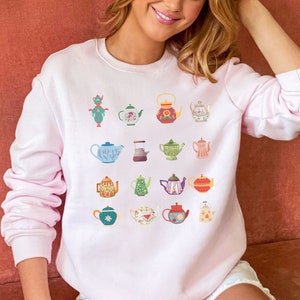 May include: A pink sweatshirt with a pattern of colorful teapots and teacups. The teapots are decorated with floral designs and have different shapes and sizes.