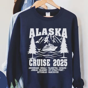以下が含まれることがあります： 白のテキストとグラフィックが特徴のネイビーブルーのスウェットシャツ。「ALASKA」の文字が山とクルーズ船のイラストの上にあり、「CRUISE 2025」が下にあります。アラスカの都市が追加のテキストでリストされています。カジュアルな旅行をテーマにした衣服。