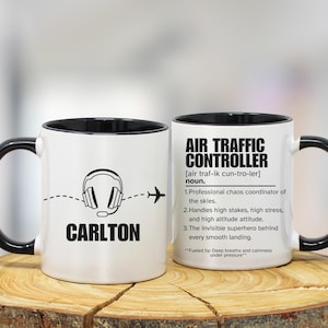 May include: Two white ceramic mugs with black handles and rims. One mug features a headset graphic, a dotted line, an airplane, and the name "CARLTON". The other mug has "AIR TRAFFIC CONTROLLER" text and a definition.