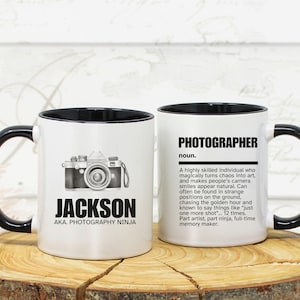 May include: Two white ceramic mugs with black handles and rims. One mug features a camera graphic and the name "JACKSON" with "AKA. PHOTOGRAPHY NINJA." The other mug has the definition of "PHOTOGRAPHER."