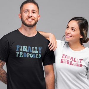 Peut inclure: Deux personnes portant des t-shirts assortis avec un design de police rétro. La personne à gauche porte un t-shirt noir avec le texte "FINALLY PROPOSED" en bleu et vert. La personne à droite porte un t-shirt blanc avec le texte "FINALLY ENGAGED" en rose et rouge.