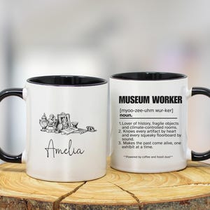 May include: Two white ceramic mugs with black handles and rims. One mug features a black and white illustration and the name "Amelia". The other mug has the text "MUSEUM WORKER" and a definition.