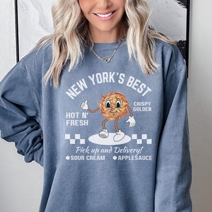 Pu&ograve; includere: Una felpa blu con un design grafico bianco che dice "New York's Best" con un'immagine di cartone animato di una patata dorata croccante. Il testo dice anche "Hot N' Fresh" e "Pick up and Delivery!" con le opzioni "Sour Cream" e "Applesauce".
