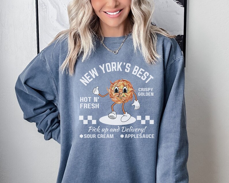 Pu&ograve; includere: Una felpa blu con un design grafico bianco che dice "New York's Best" con un'immagine di cartone animato di una patata dorata croccante. Il testo dice anche "Hot N' Fresh" e "Pick up and Delivery!" con le opzioni "Sour Cream" e "Applesauce".