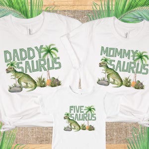 Op de afbeelding: Witte T-shirts met een dinosaurus- en palmboommotief. De shirts zeggen "Daddy Saurus", "Mommy Saurus" en "Five Saurus".