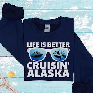 以下が含まれることがあります： 「LIFE IS BETTER CRUISIN' ALASKA」の文字が入ったネイビーブルーのスウェットシャツ。クルーズ船と山の風景が描かれたサングラスのデザイン。スウェットシャツは長袖です。