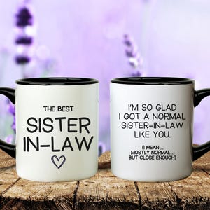 May include: Two white ceramic mugs with black handles and rims. One mug reads "THE BEST SISTER IN-LAW" with a heart. The other mug says "I'M SO GLAD I GOT A NORMAL SISTER-IN-LAW LIKE YOU."